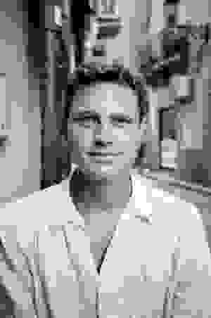 Bild von Raphael Volkmer in schwarz weiß. Junger kurzhaariger blonder Mann mit weißen Hemd, im Hintergrund ist eine südländisch anmutende Straße zu sehen