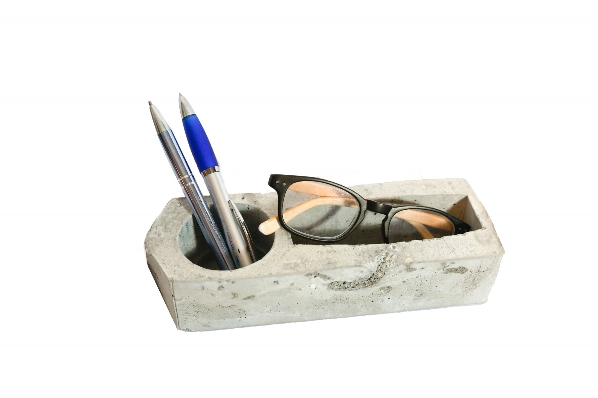 Ein Stiftehalter aus Beton mit zwei Fächern. In einem stecken zwei Kugelschreiber und im anderen liegt eine Brille.
