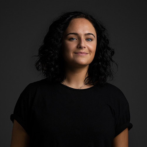 Portrait einer lächelnden jungen Freelancerin mit schwarzen mittellangen Locken vor schwarzem Hintergrund